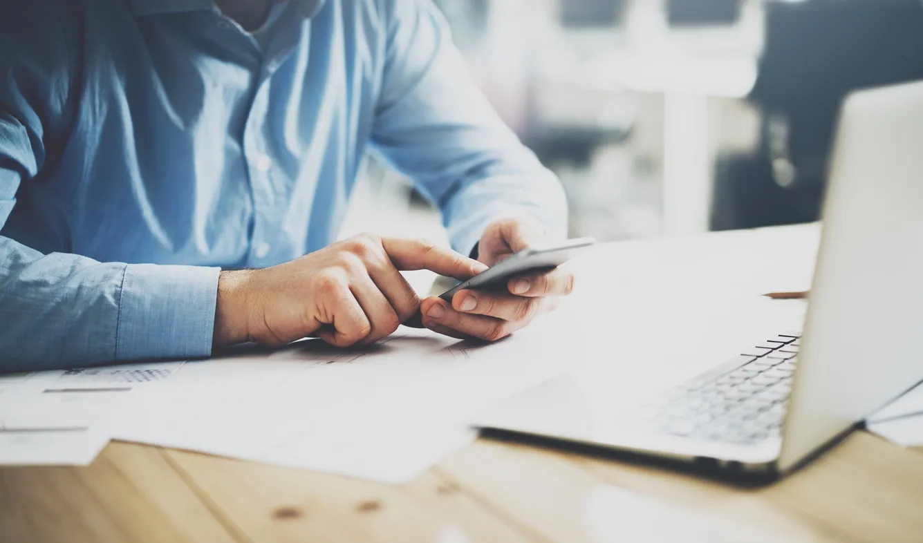 Person using smartphone at desk with laptop and papers.